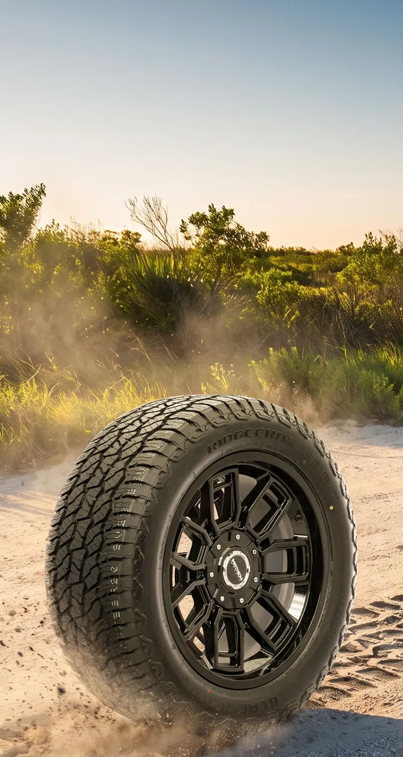 Blackhawk Ridgecrawler all-terrain tire mounted on a black wheel kicking up sand on an off-road trail, surrounded by green shrubs under a clear blue sky.